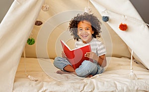 Cute little african american kid curly boy reading book in play tent