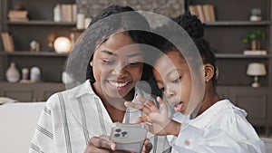 Cute little african kid child daughter having fun with multiracial mum at home using smart phone typing swipe screen