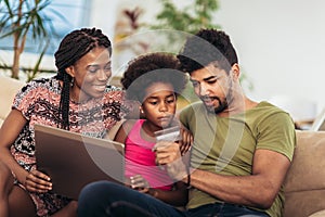 Cute little Afro-American girl and her beautiful young parents using laptop