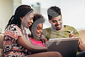 Cute little Afro-American girl and her beautiful young parents using a laptop and doing shopping online