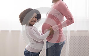 Cute little afro girl hugging her pregnant mom`s tummy and smiling