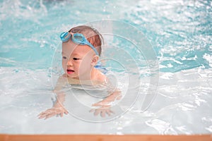 Cute little Asian 18 months toddler boy child in trunks wear swimming goggles learn to swim at indoor pool