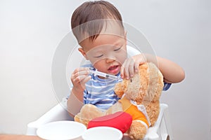 Cute little Asian 2 years old toddler baby boy child playing feeding teddy bear with liquid medicine with a syringe at home