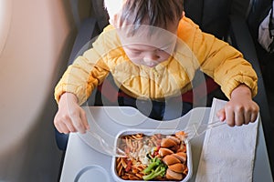 Cute little Asian 2 years old toddler baby boy child wearing yellow jacket eating food pasta during flight on airplane