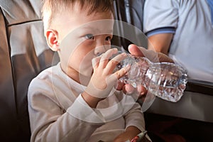 Cute little Asian 3 years old toddler boy child drinking water from bottle during flight on airplane. Little kid feeling earache