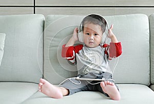 Cute little Asian baby boy in headphones is using a smartphone lying on the sofa at home. Child listening to music on earphones