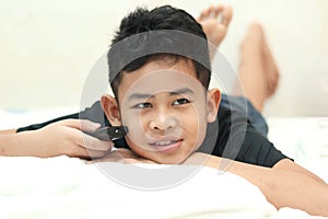 Cute little Asian boy child lying in bed holding the tv remote control and watching television in bedroom at home