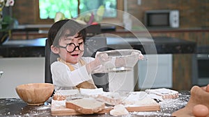 Cute little Asian boy sifting dough flour with sifter sieve colander in home kitchen for prepare to baking bakery and cake. Thai k