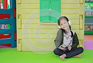 Cute little Asian child girl listening music by headphone near toy playhouse in playground