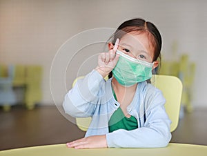 Cute little Asian child girl wearing a protective mask with showing one forefinger sitting on kid chair in children room