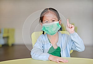 Cute little Asian child girl wearing a protective mask with showing one forefinger sitting on kid chair in children room