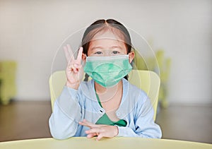 Cute little Asian child girl wearing a protective mask with showing three fingers sitting on kid chair in children room