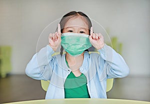 Cute little Asian child girl wearing a protective mask with showing two forefingers sitting on kid chair in children room