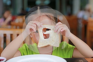 Cute little asian girl looking through hole of bite bread sheet. Asian girl having breakfast. Child looking at camera