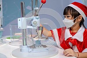 Cute little Asian girl in red santa dress looking at microscope in classroom. Education science project concept