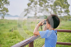 Cute little Asian school boy child looking through binoculars explores nature field at viewpoint outdoors