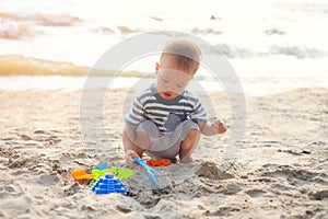 Cute little Asian toddler boy sitting & playing children`s beach toys  on beautiful sandy tropical sunset beach