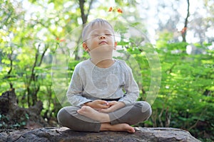 Cute little Asian 2 - 3 years old toddler baby boy child with eyes closed, barefoot practices yoga & meditating outdoors on nature