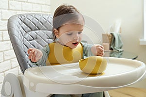 Cute little baby with bowl sitting in high chair indoors