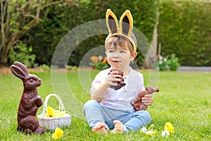 Cute little baby boy with bunny ears eating chocolate Easter bunnies sitting on green grass outside in the spring garden with bask