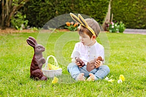 Cute little baby boy with bunny ears eating chocolate Easter bunnies sitting on green grass outside in the spring garden