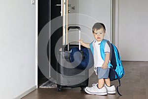 Cute little baby boy in fathers shoes and with big backpack holding suitcase staying near open door ready to travel leaving home.