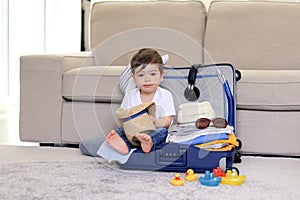 Cute little baby boy with funny face expression sitting in blue suitcase holding straw hat in hands, packed for vacation,