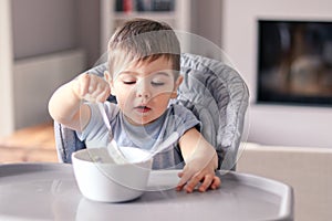 Cute little baby boy with funny smeared face concentrated on food eating with fork from white bowl at table in front of him sittin