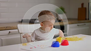 Cute little baby boy sitting behind table and drawing with fingers covered in colorufl paint. Concept of child education