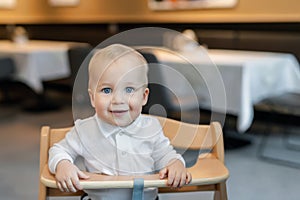 Cute little baby boy in white polo t-shirt sitting in wooden baby chair and laughing at cafe indoors. Portrait of