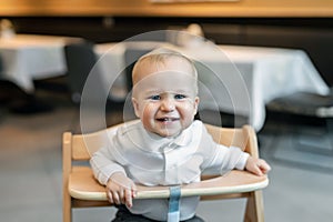 Cute little baby boy in white polo t-shirt sitting in wooden baby chair and laughing at cafe indoors. Portrait of