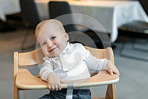 Cute little baby boy in white polo t-shirt sitting in wooden baby chair and laughing at cafe indoors. Portrait of