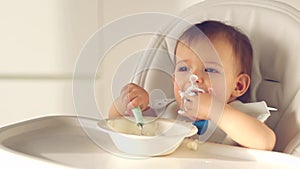 Cute little baby girl eating baby food with spoon. her face is smeared with yoghurt on high chair in home kitchen