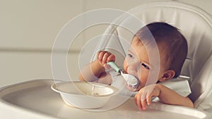 Cute little baby girl eating baby food with spoon on high chair in home kitchen