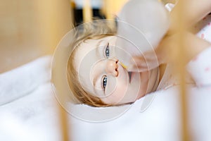 Cute little baby girl holding bottle with formula mild and drinking. Child in baby cot bed before sleeping