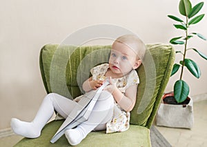 Cute little baby girl make funny face, holding book in hand. Kid sitting on armchair.