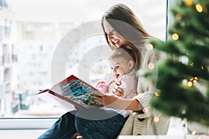 Cute little baby girl in pink dress with her mother young woman reading book in room at home, Christmas time at home, happy New
