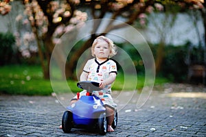 Cute little baby girl playing with blue small toy car in garden of home or nursery. Adorable beautiful toddler child