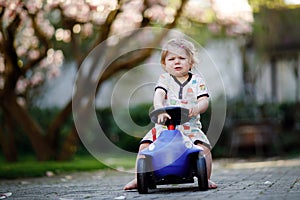 Cute little baby girl playing with blue small toy car in garden of home or nursery. Upset crying toddler child outdoors.