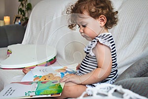 Cute little baby girl reading a picture book on a white sofa
