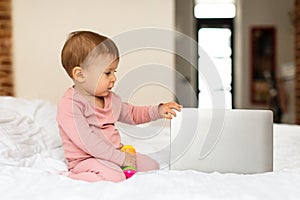 Cute little baby girl sitting on bed and touching laptop computer, bedroom interior, free copy space