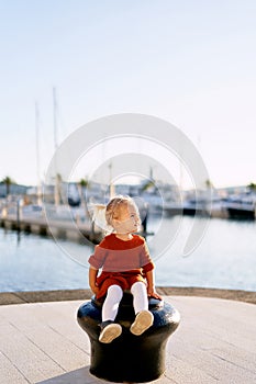 Cute little baby girl in a terracotta dress and white tights is sitting on a boat pier by the sea