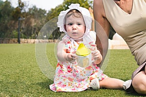 A cute little baby girl toddler wearing a white bonnet and a dress sat on green grass with her grandmother and holding a sippy cup