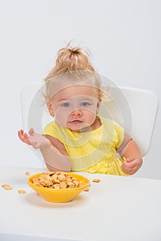 Cute little baby girl 1 year old in yellow t-shirt eating cereal flakes at the table isolated on white background