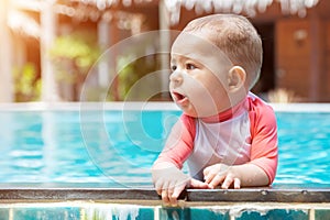 A cute little baby holds to the edge of the tropical pool and looks to the left. Infant girl swim at summer vacation