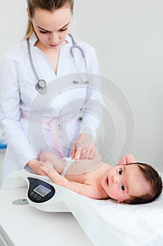 Cute little baby lying on scales, medical checkup in clinic, copy space. Young female blonde pediatrician doctor examines baby