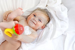 Cute little baby playing with toy rattle and own feet after taking bath. Adorable beautiful girl wrapped in white towels