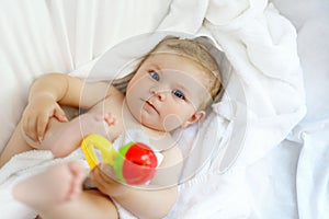 Cute little baby playing with toy rattle and own feet after taking bath. Adorable beautiful girl wrapped in white towels