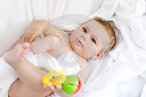 Cute little baby playing with toy rattle and own feet after taking bath. Adorable beautiful girl wrapped in white towels