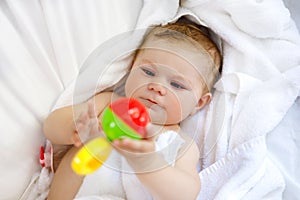 Cute little baby playing with toy rattle and own feet after taking bath. Adorable beautiful girl wrapped in white towels